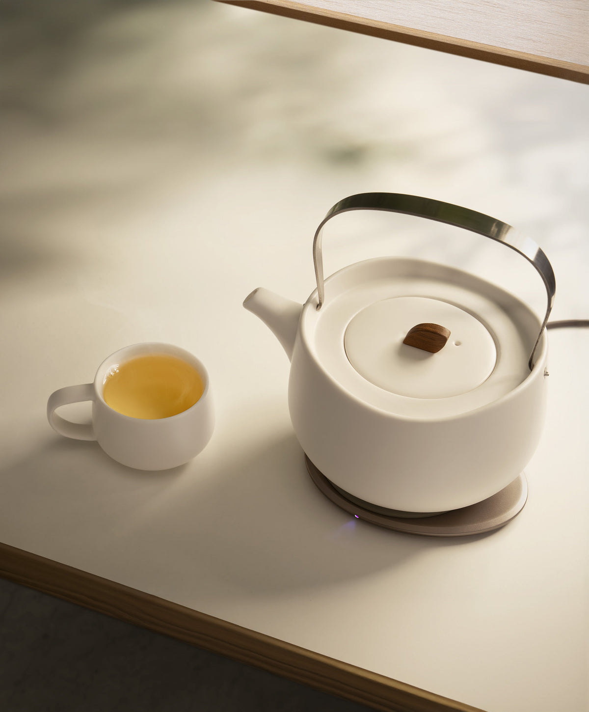Top view of Leiph self-heating ceramic teapot 20 fl oz / 600 mL in Jasmine White charging on OHOM Pad 3 with tea in teacup beside it