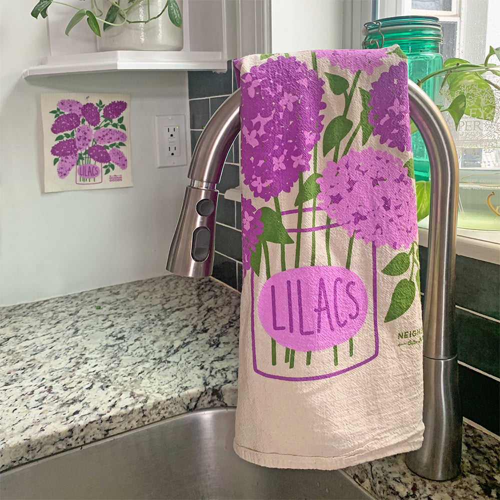The Neighborgoods Lilacs dish towel with a lilac flower illustration hanging over a kitchen sink faucet.