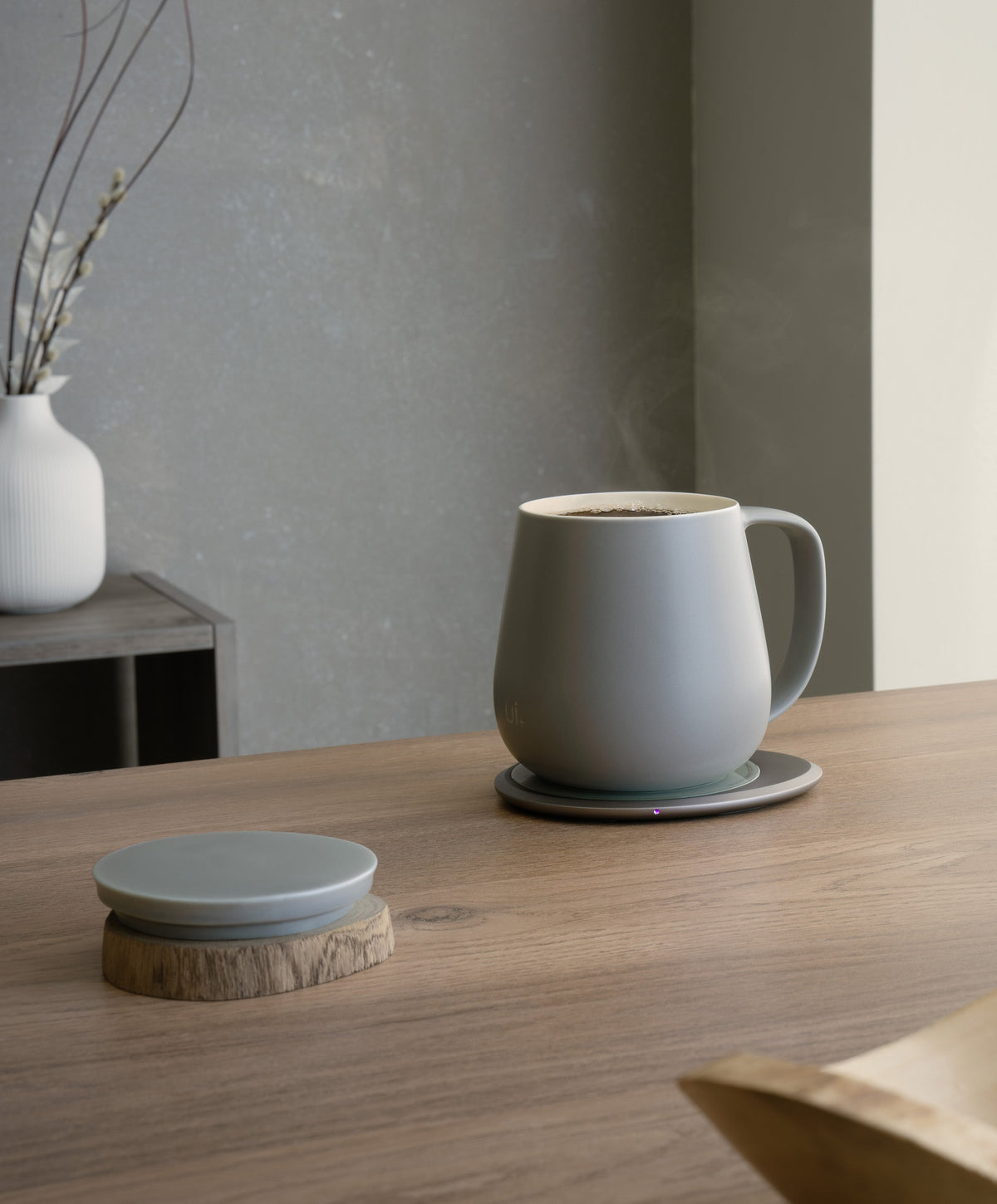 Ui+ self-heating ceramic mug set 18 fl oz / 532 mL in Soft Gray charging on OHOM Pad 3, lid placed on wooden coaster
