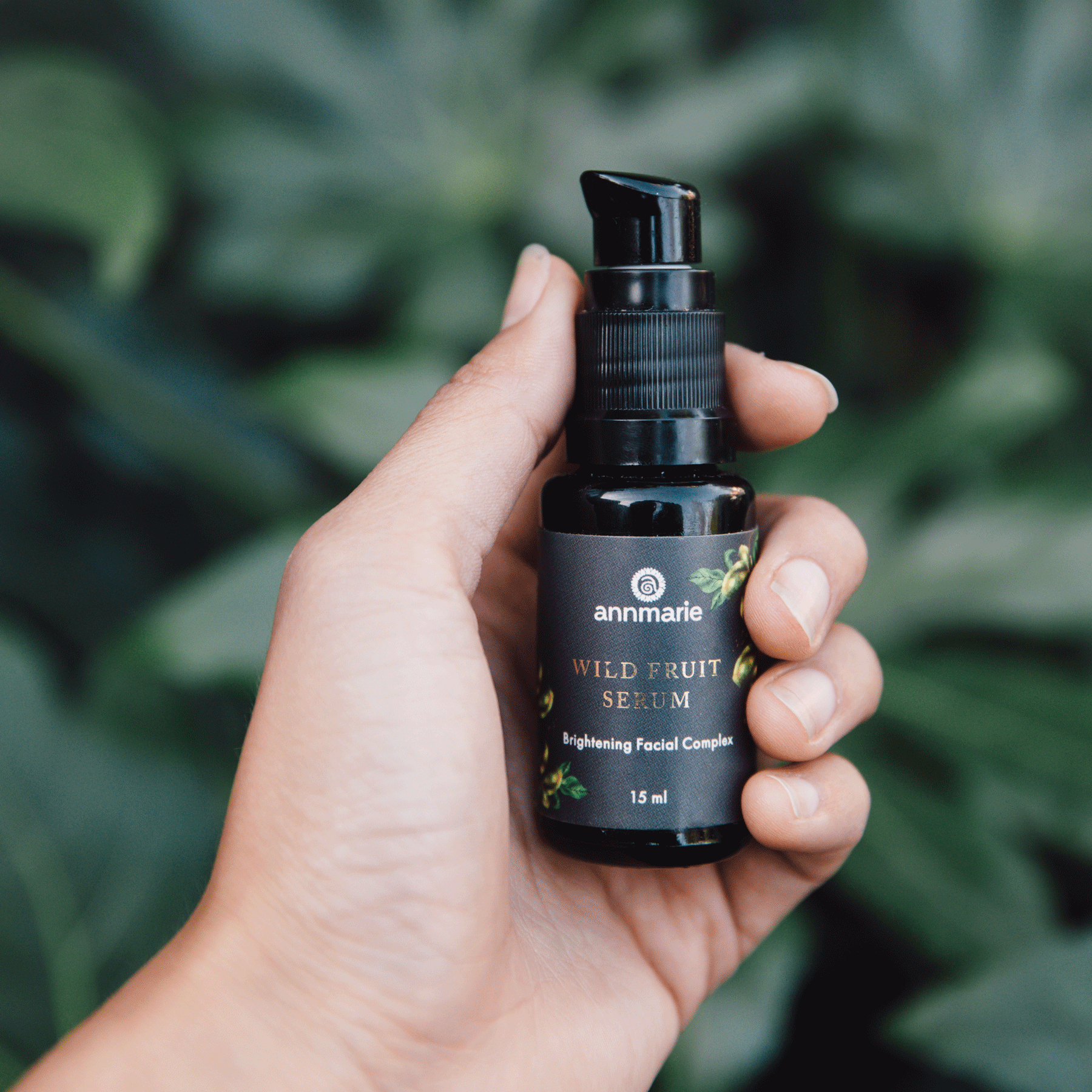 Hand holding Annmarie Wild Fruit Serum brightening facial complex in a small pump bottle against a green natural background.