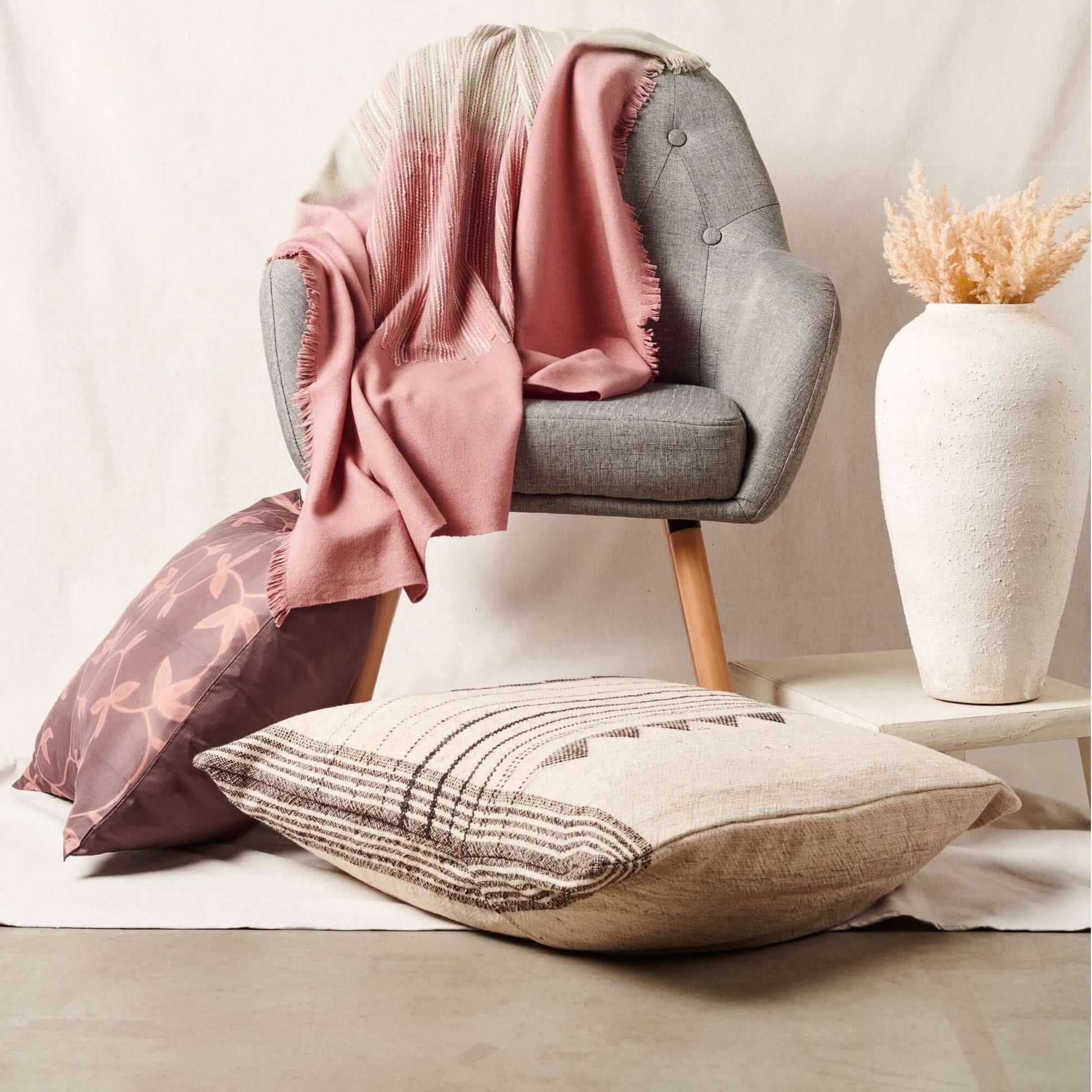 Chive Pink Green Ombre Handloom Merino Throw draped over a gray chair with decorative pillows and a vase.