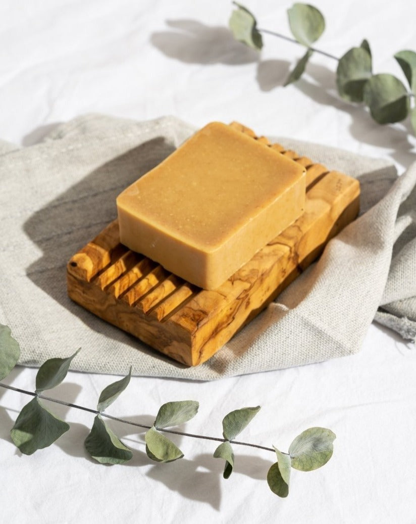 Olive wood soap holder with soap placed on top, flanked by decorative stems with leaves, and a cloth underneath the holder