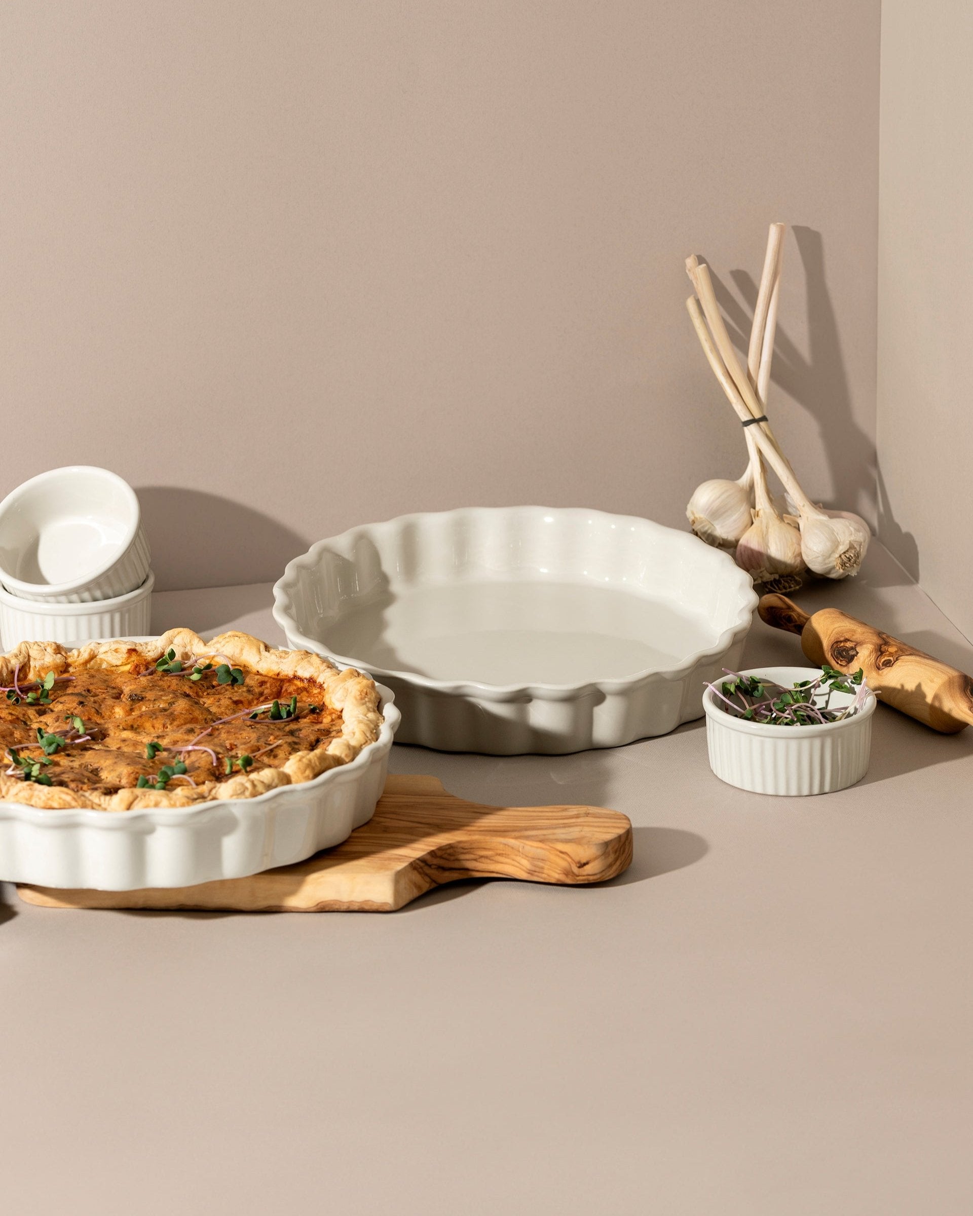A white rounded stoneware pie dish with wavy side walls displayed on a neutral background, accompanied by a wooden cutting board with a pie on it, some garlic bulbs, and a small white bowl.