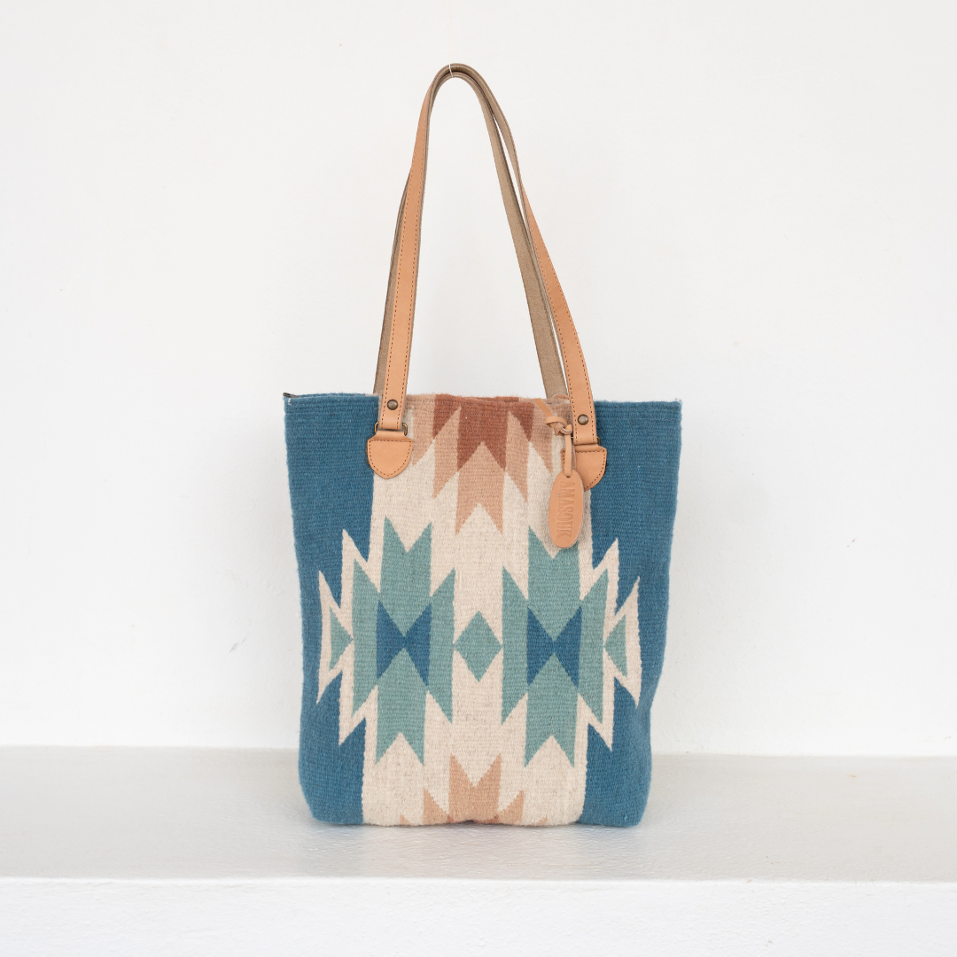 Handwoven bucket bag in rose and blue with abstract diamond and lightning pattern, leather shoulder straps, and relaxed shape, ideal for everyday or beach use.