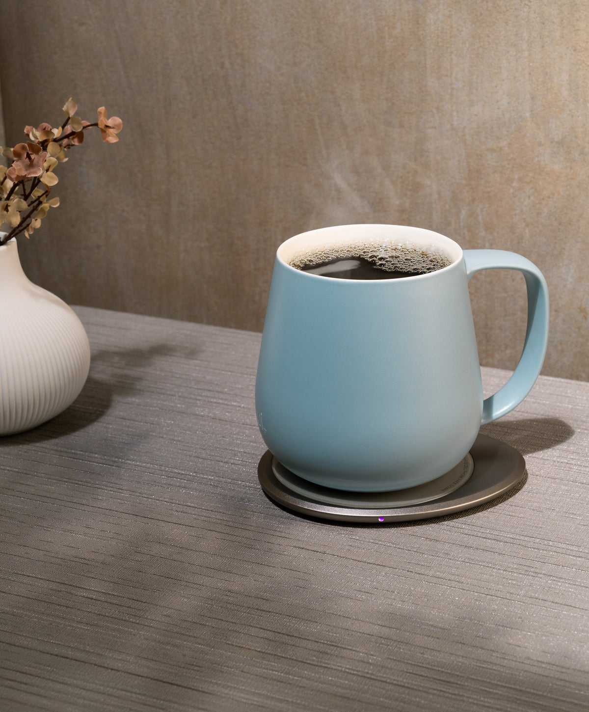 Ui+ self-heating ceramic mug set 18 fl oz / 532 mL in Moonmist Blue charging on OHOM Pad 3, filled with hot coffee and steam rising