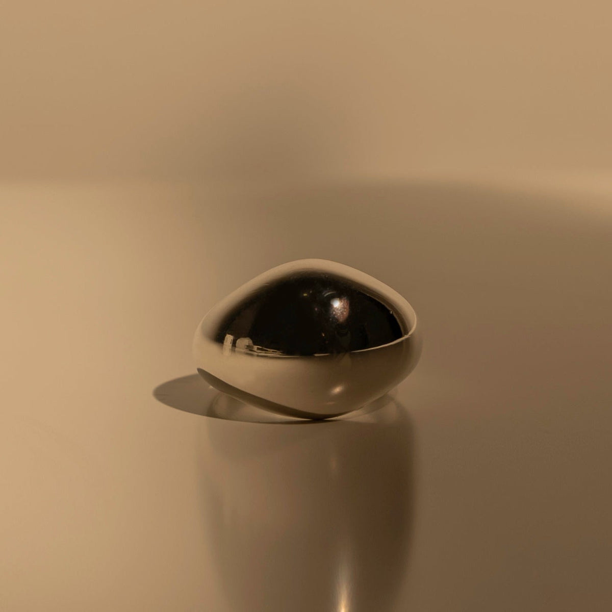 A minimalist dome-shaped ring with a polished silver-toned outer band and a dark reflective center, displayed on a smooth reflective surface under warm, dim lighting.