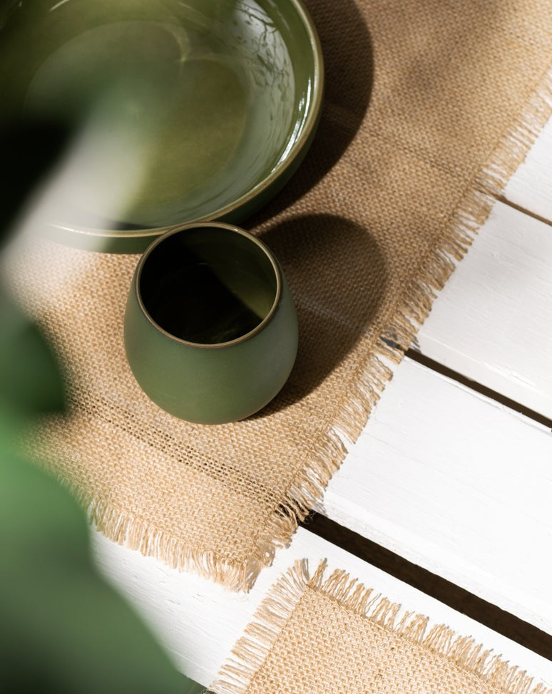 A beige burlap placemat on a gray surface, featuring an empty green plate and a matching green cup, arranged for a clean and natural table setting.