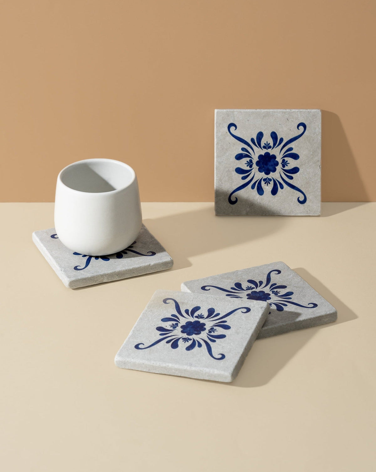 A set of four square white marble coasters with blue floral patterns, displayed with a cup on one of them, on a beige background.