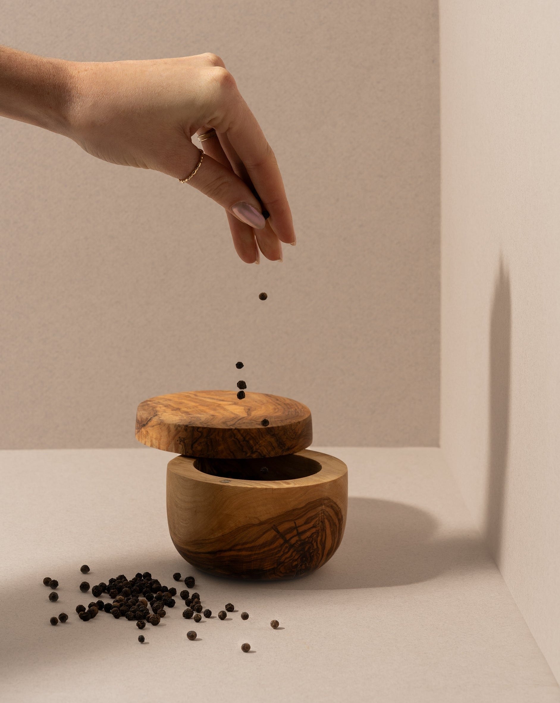 Hand sprinkling spices over a partially covered round olive wood salt and spice cellar, set against an off-white background.