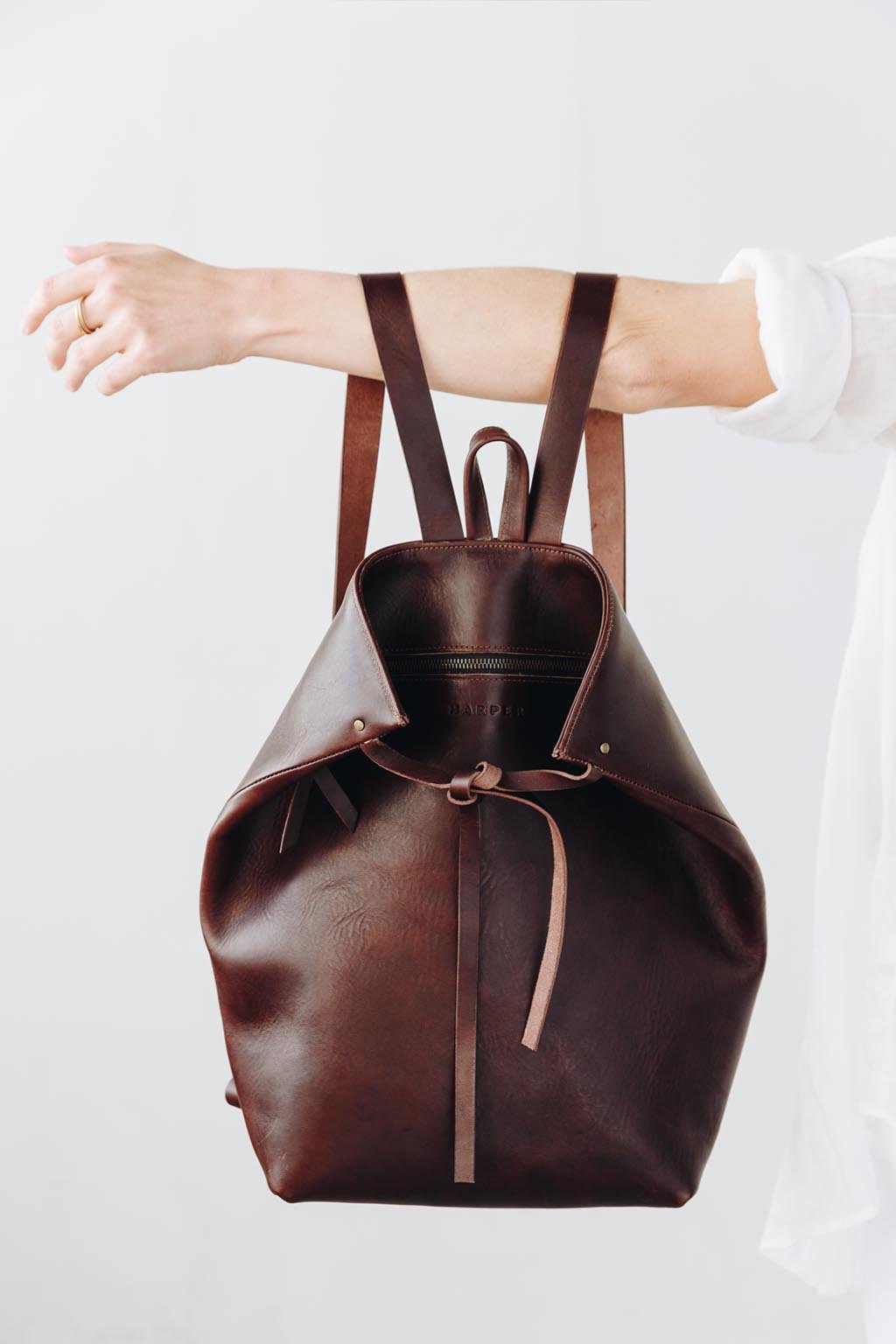A woman holds out her arm, which is looped through the shoulder straps of a dark brown leather backpack. The top corners of the backpack are cinched with straps that knot over a zipper, creating a unique shape.