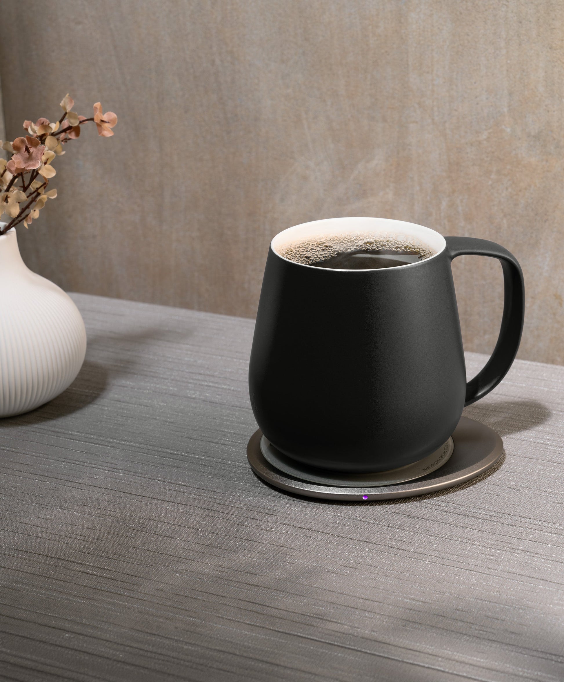 Ui+ self-heating ceramic mug set 18 fl oz / 532 mL in Inkstone Black charging on OHOM Pad 3, filled with hot coffee and steam rising