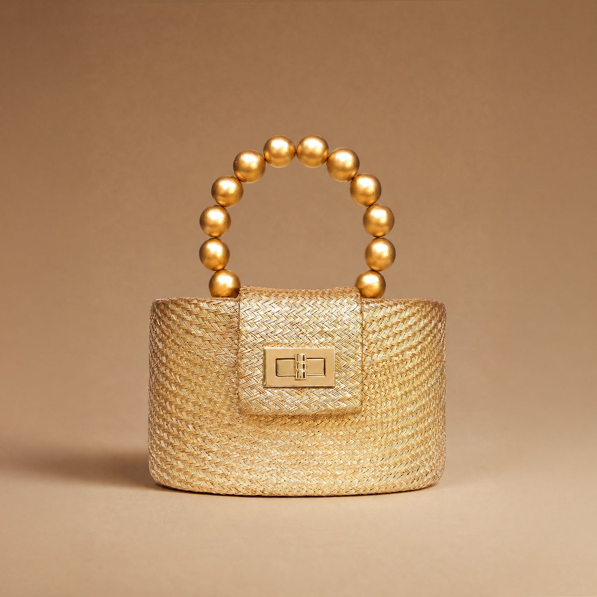 A small structured handbag in woven straw with a gold-tone turn-lock clasp and a curved top handle made of glossy gold beads, photographed on a warm beige studio background.