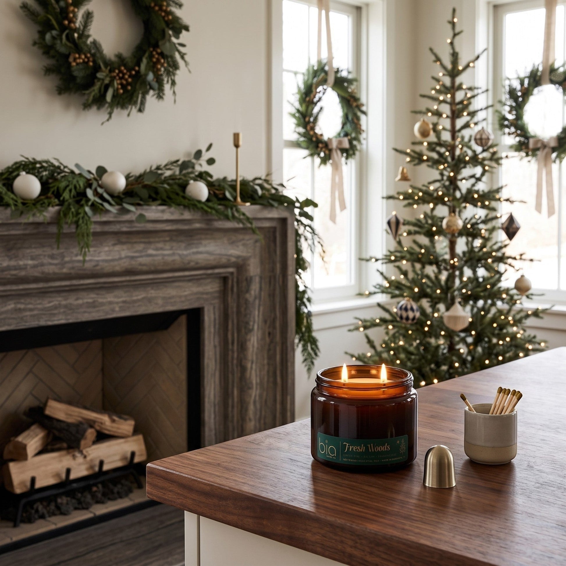 A lit amber glass jar candle sits on a wooden kitchen counter in a decorated holiday home, with a fireplace, wreaths, and a Christmas tree in the background.