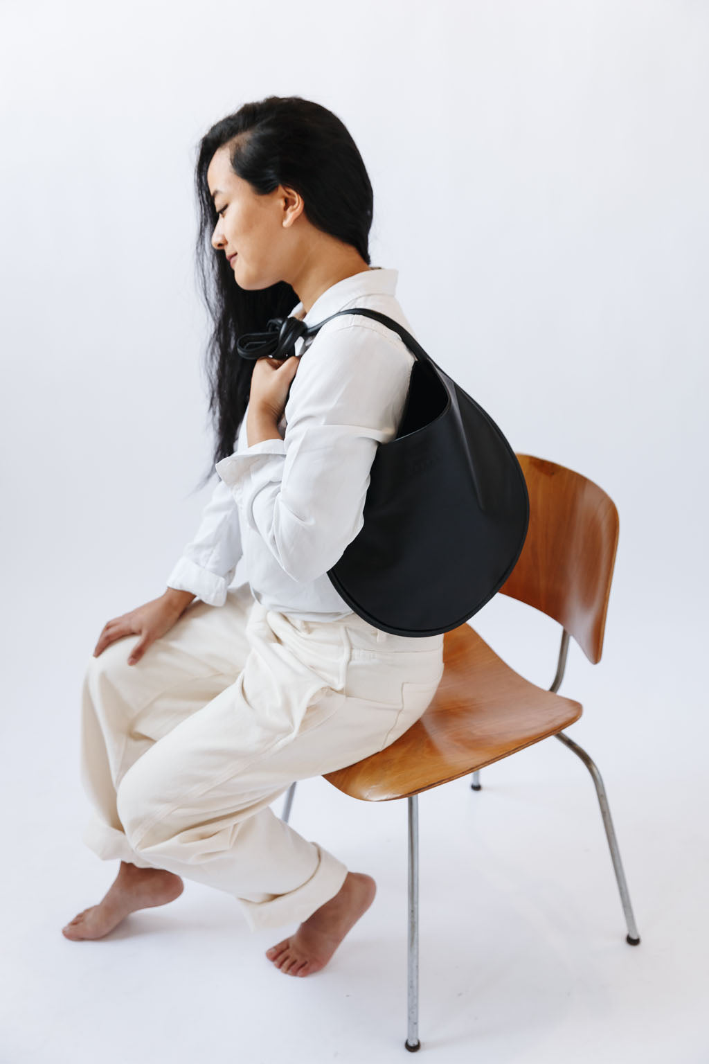 A woman sits on a chair, wearing a black leather shoulder bag over her shoulder. The shoulder strap is tied so a knot sits over her shoulder. The leather is soft and shiny, and the bag is a half moon shape.
