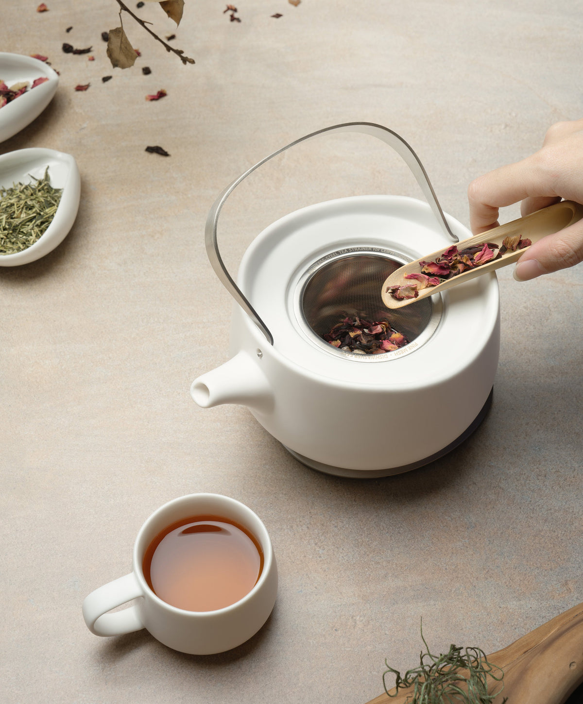 Person dropping tea leaves into Leiph self-heating ceramic teapot 20 fl oz / 600 mL in Jasmine White with brewed tea in teacup beside it