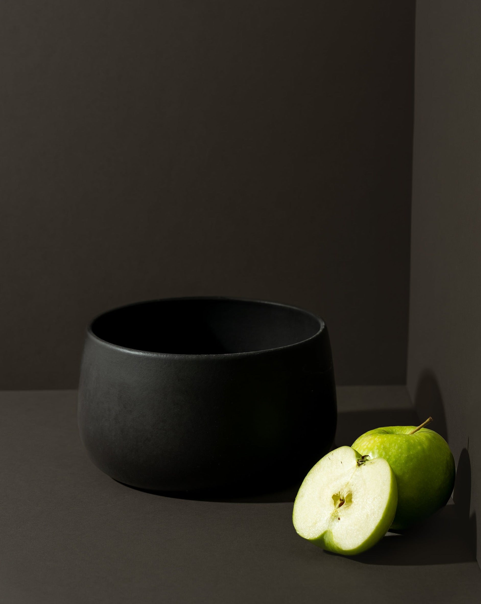 A person is serving salad from a black stoneware serving bowl on a dining table, with a white plate and a glass of white wine in the background.