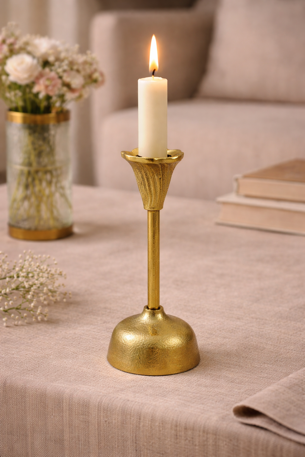 Gold candle holder with a lit candle on a table with a vase of flowers and books in the background.
