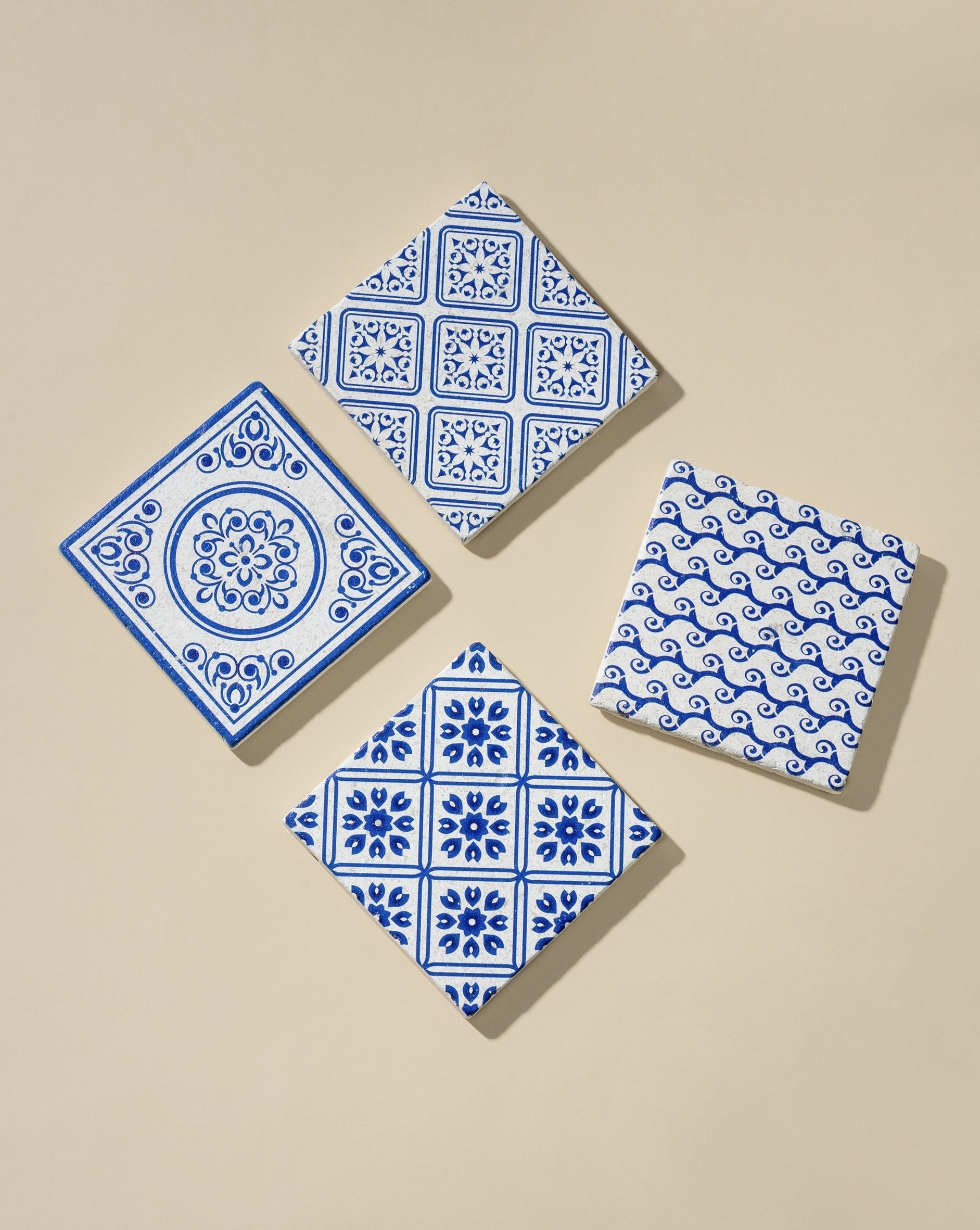 Four square white marble coasters with blue patterned designs, arranged flat on an off-white background, highlighting their hand-finished details and elegant look.
