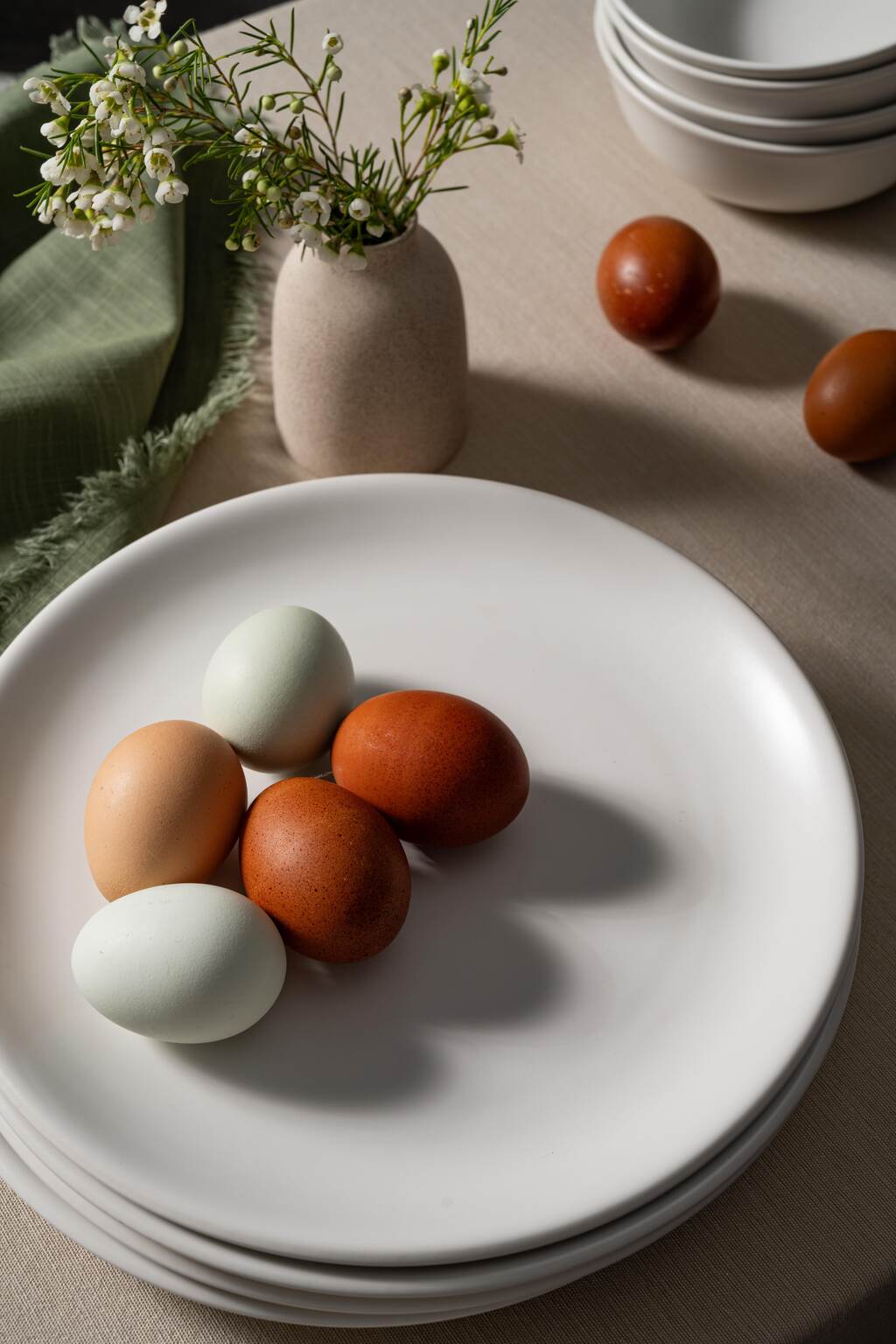 Bone-colored ceramic dinner plates from Hamkke Home with multicolored farm eggs, styled with a small vase and stacked bowls on a neutral table setting.