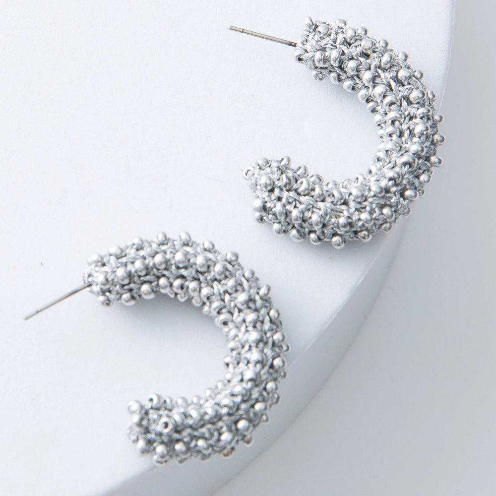 These are silver demi-hoop earrings that are open at the back. They are composed of silver thread and silver beads crocheted together to form a chunky hoop. 