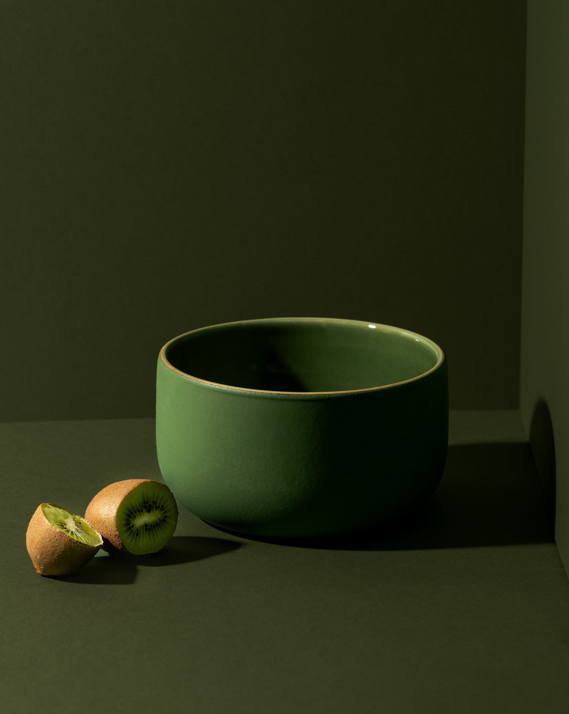 Matte green stoneware bowl displayed against a green background, with two slices of kiwi beside it