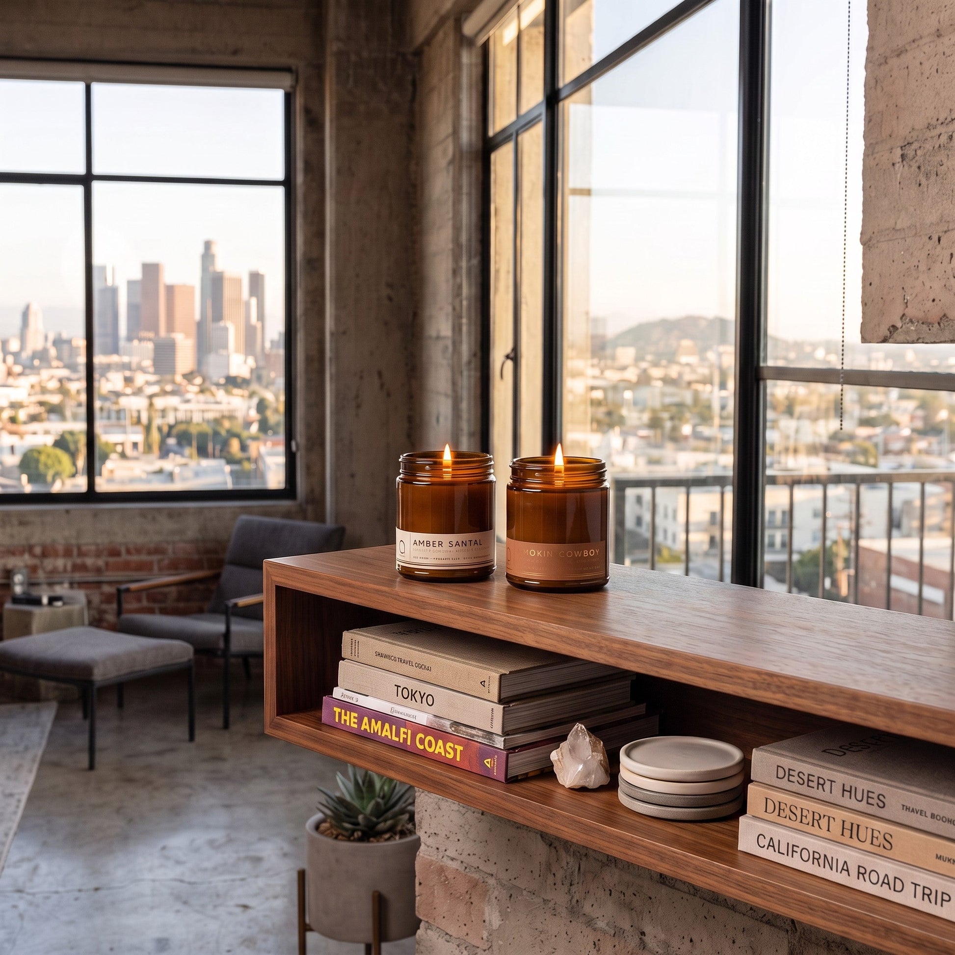 Two amber glass jar candles labeled 'LAUREL & VANILLA' are lit on a wooden floating shelf in a loft-style room with a city skyline view outside.