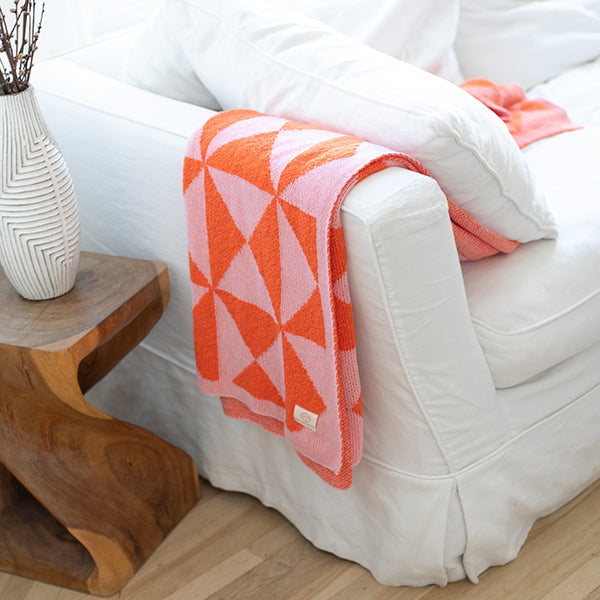 White sofa with a pink and orange windmill patterned blanket draped over it, next to a wooden side table with a decorative vase.