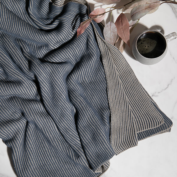 Marine blue and gray textured blanket on a white surface with a cup of coffee and decorative leaves.