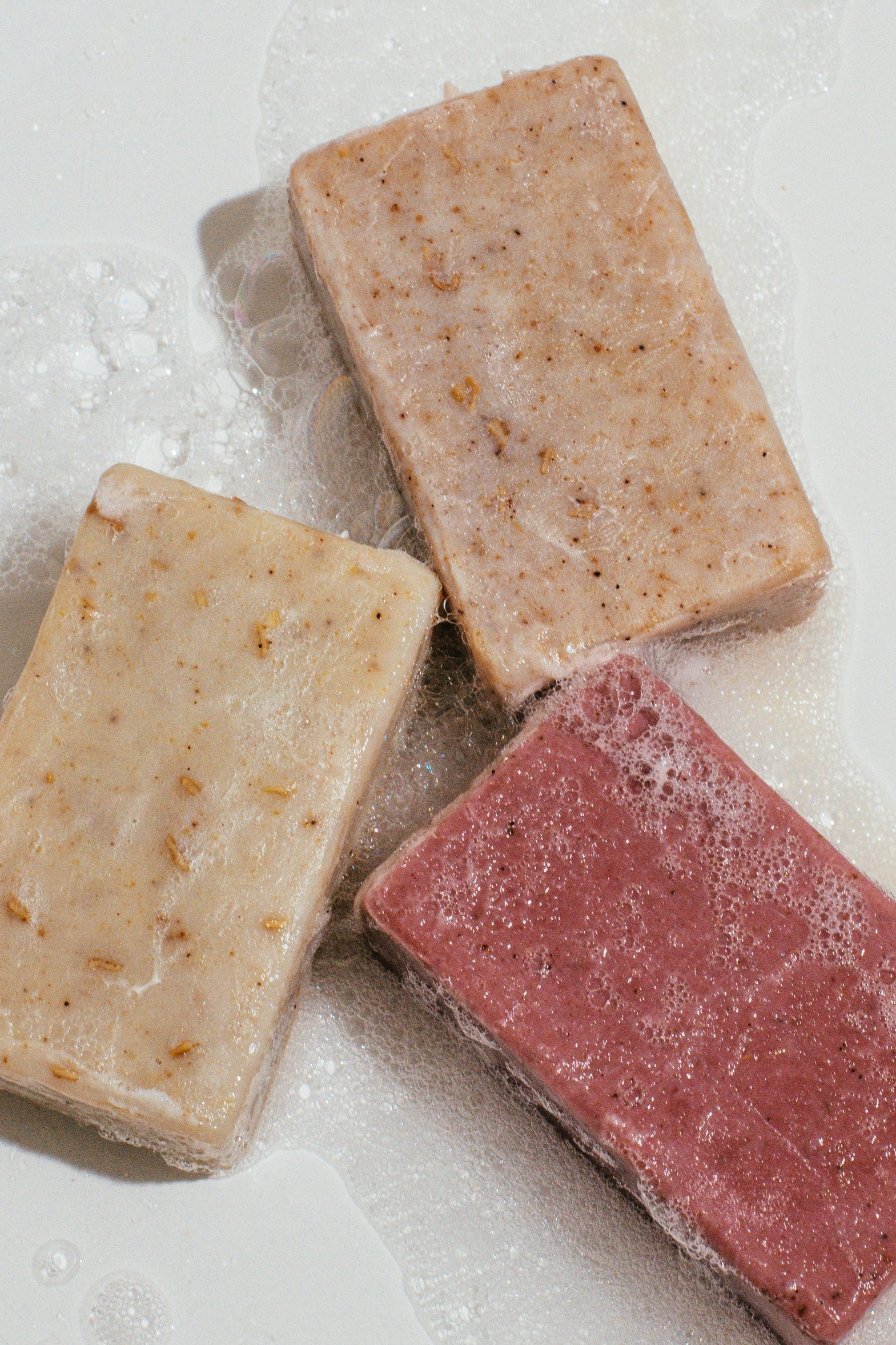 Close up of Wenjulan Whole Body Care Cleansing Bar Trio bundle gift pack featuring Wenjulan Unscented Cleansing Soap with Colloidal Oatmeal bar, Wenjulan Sensitive Skin Cleansing Soap with Kaolin Clay bar, Wenjulan Southwest Cedar Cleansing Soap bar with suds and lather on a light gray background
