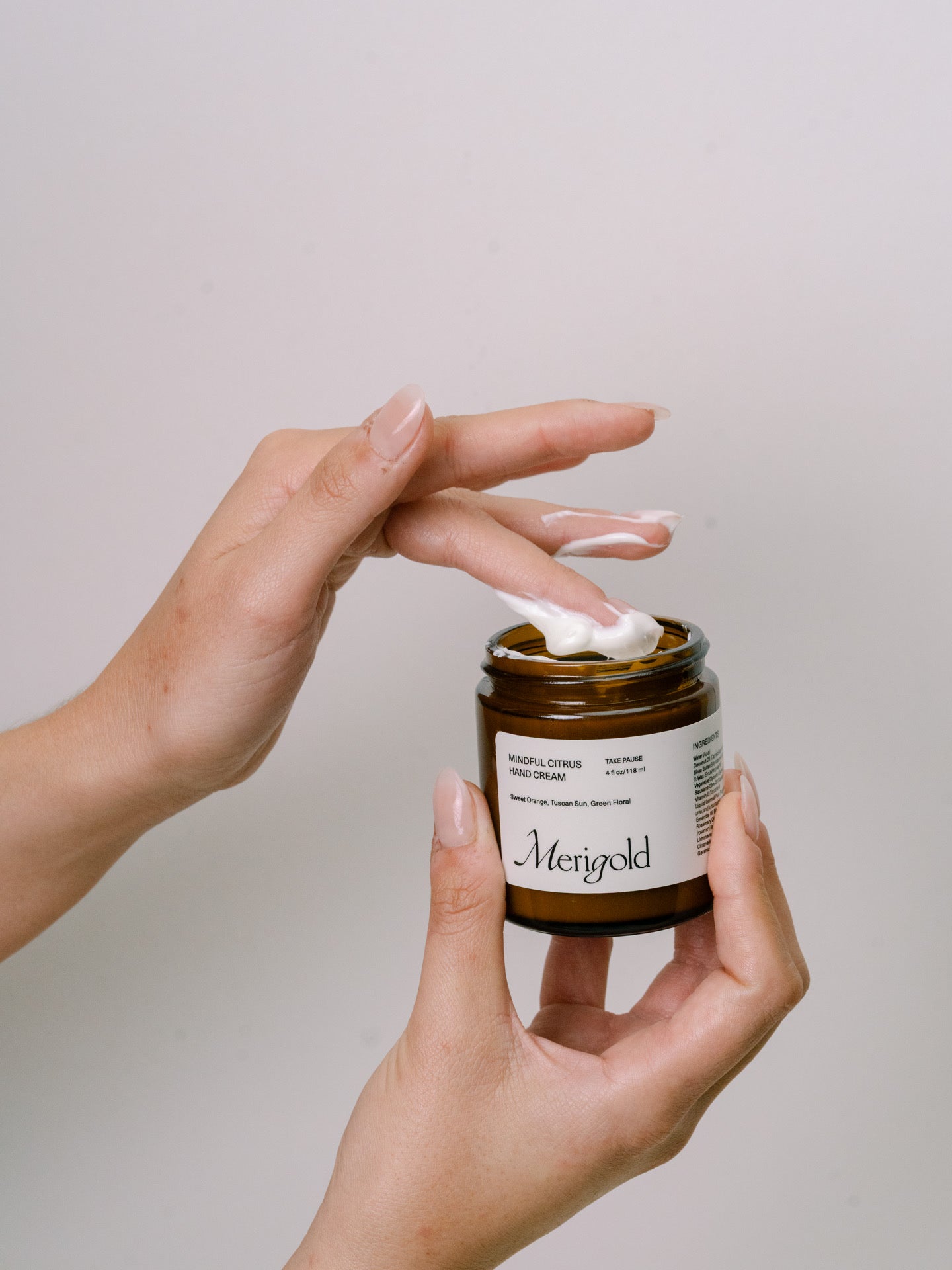 Hand holding a jar of Merigold Mindful Citrus hand cream, dipping fingers showing texture with a white background