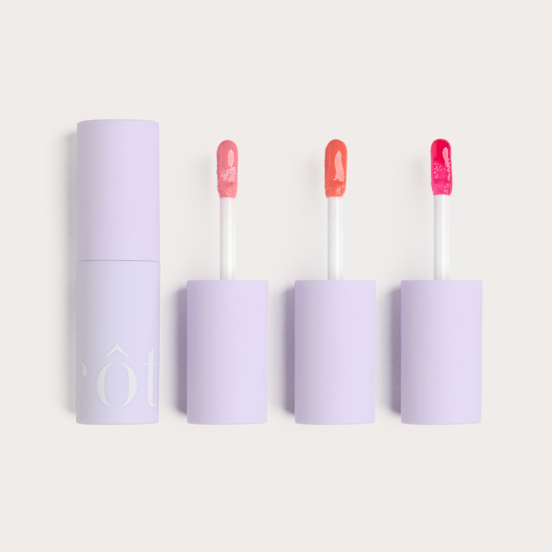 Three lip gloss containers with their respective applicators displayed side by side, with the central container slightly advanced, showcasing Côte Beauty The Darlings Lip Trio Peptide Non-Toxic Lip Gloss Set in, Light Pink, Coral and Magenta shades. 
