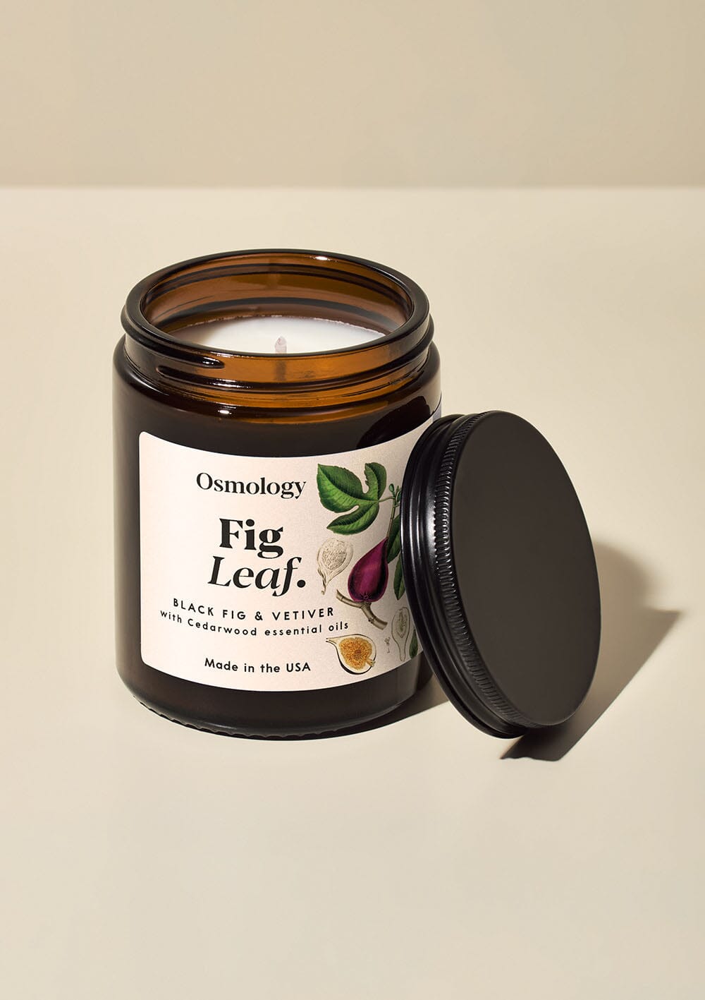 Osmology Fig Leaf jar candle, brown glass jar with white decorative label, black lid leaning against it, on white background