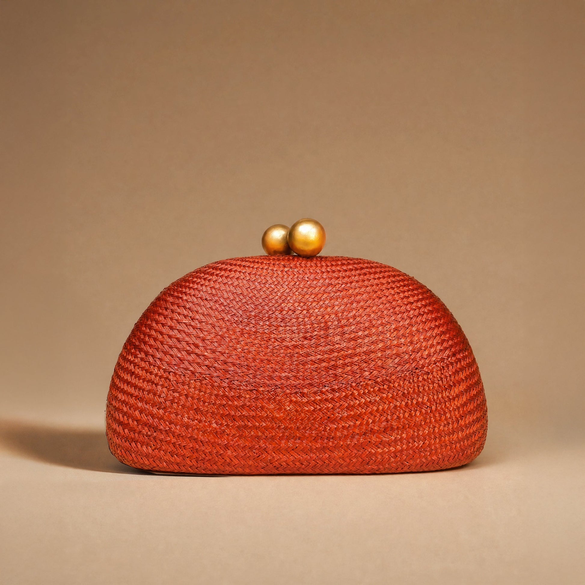 A textured woven clutch bag in a vibrant rust-orange color, featuring a domed shape and two small gold-tone pearl clasp buttons on top, set against a solid warm beige background.