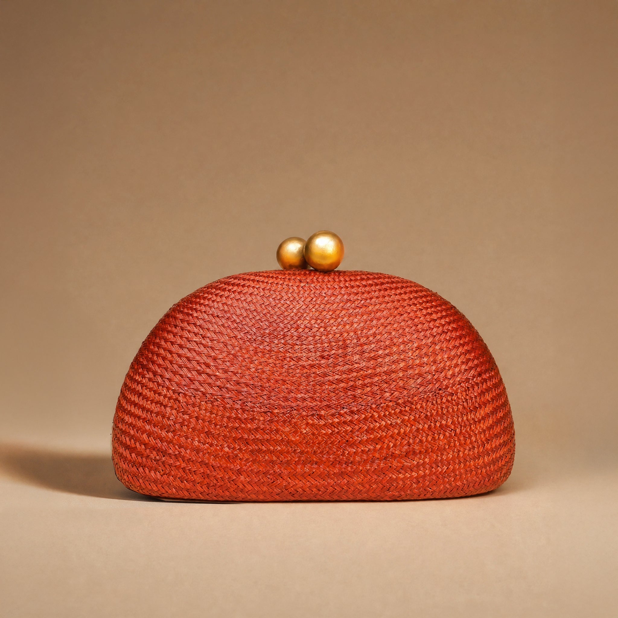 A textured woven clutch bag in a vibrant rust-orange color, featuring a domed shape and two small gold-tone pearl clasp buttons on top, set against a solid warm beige background.