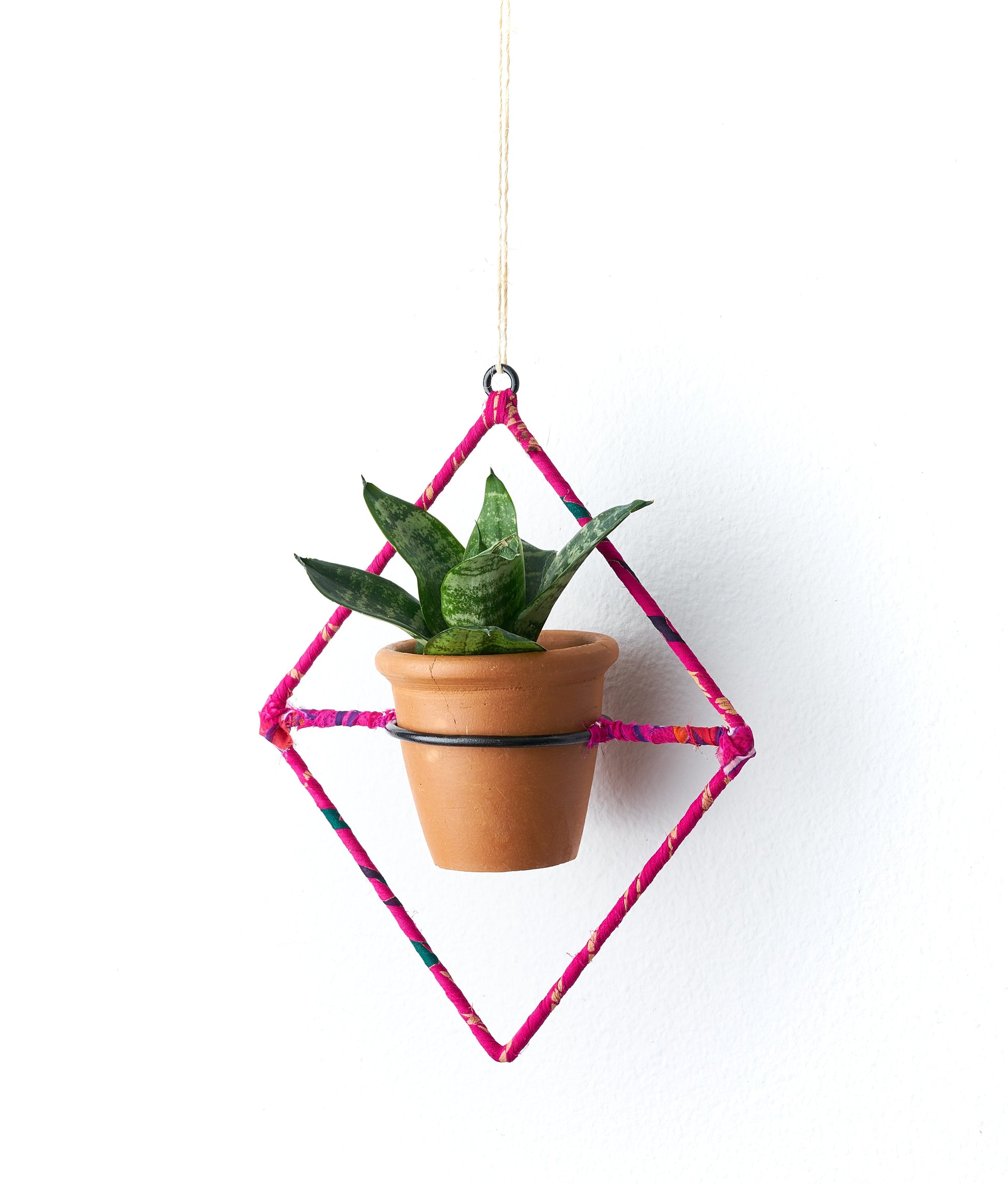 This handcrafted hanging planter showcases a small terracotta pot suspended within a diamond-shaped frame, wrapped in assorted upcycled sari fabric. It includes a drainage hole for proper water management.