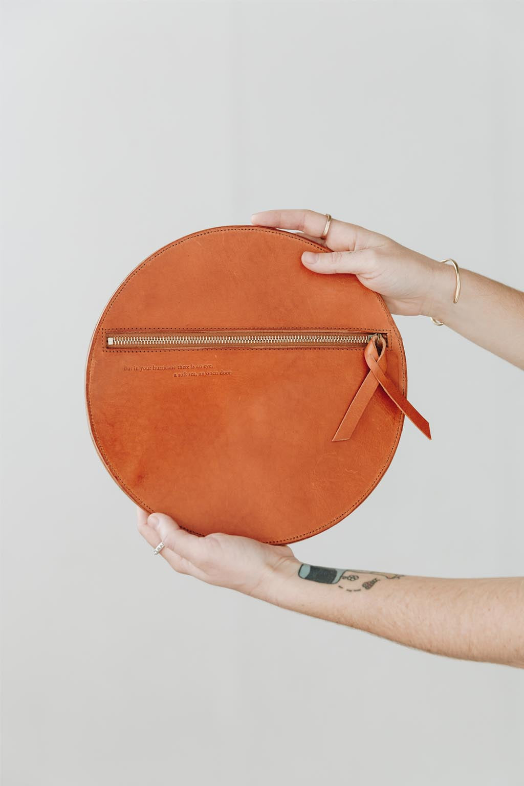 A woman's arms are visible, holding out a circular natural leather clutch with the reverse side and zipper visible. The zipper includes a matching zipper pull.