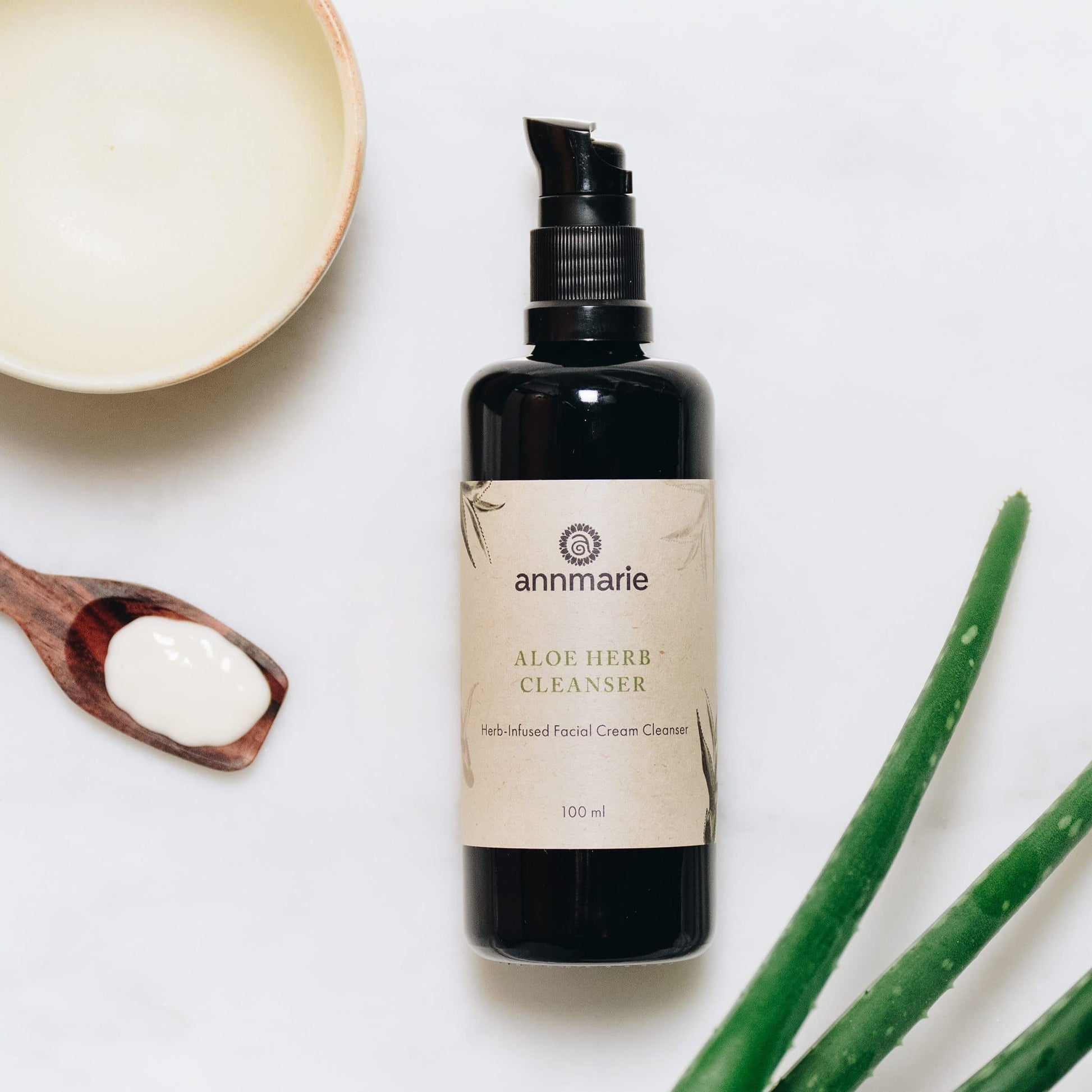 Black pump top 100 ml bottle of Annmarie Aloe Herb Cleanser with a wooden spoon and aloe vera leaves on a white background