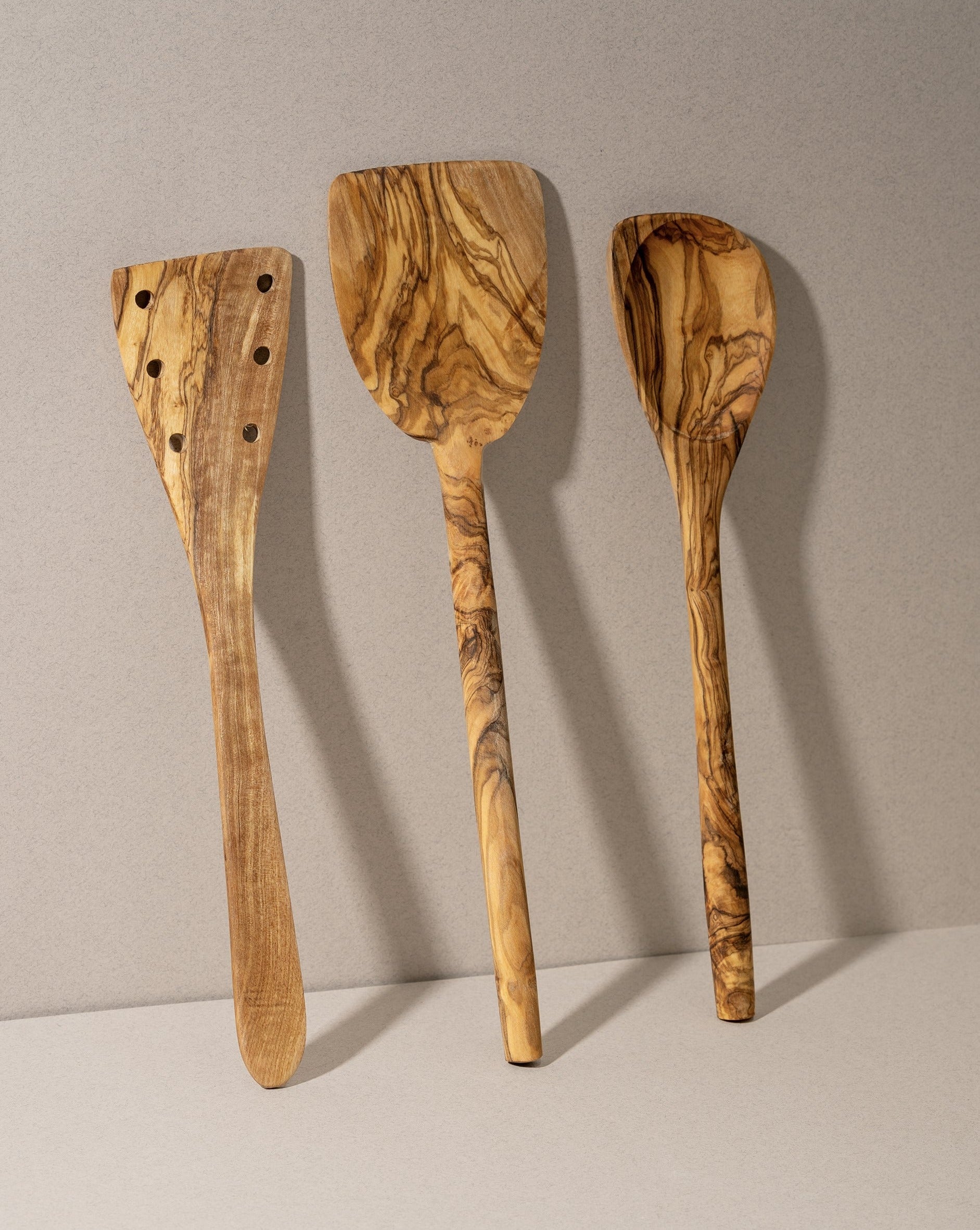 A set of three olive wood kitchen utensils, including a flipper, a pointed spoon, and a pancake spatula, laid out on a neutral surface.