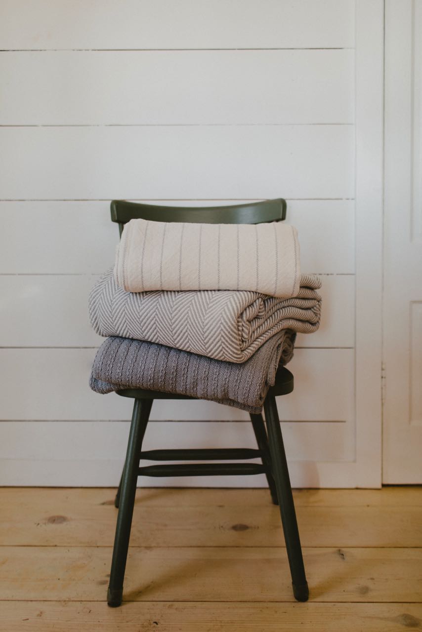  A simple display of Evangeline linens, showcasing a stack of luxurious blankets on a chair against a pristine white wall. Pinstripe patterns adorn these 100% Cotton bed blankets, available in king, queen, and twin sizes. Crafted with durability and washability in mind, these heirloom-quality blankets exude opulence.