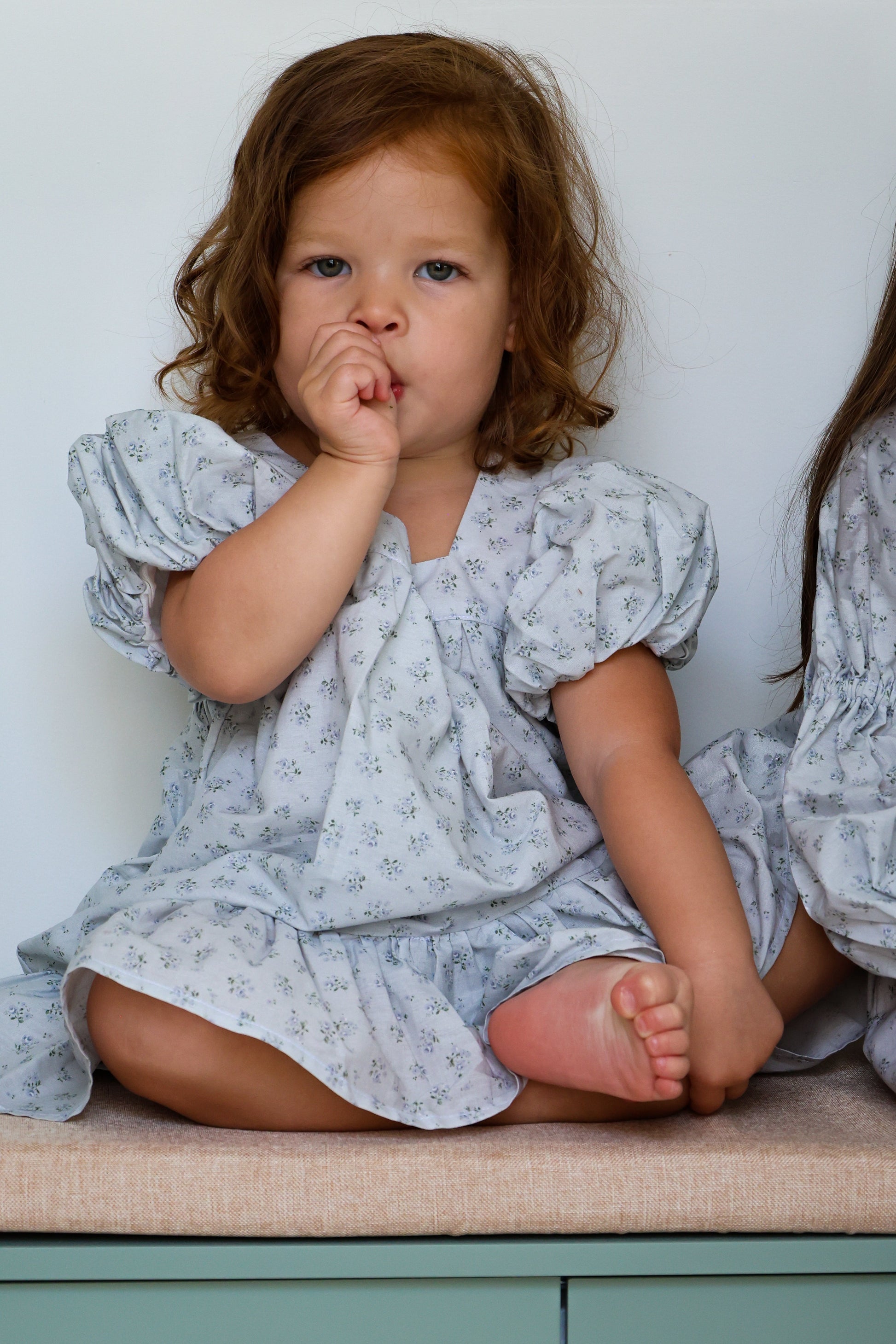 Child in a light blue floral dress sitting on a beige cushion against a white wall.