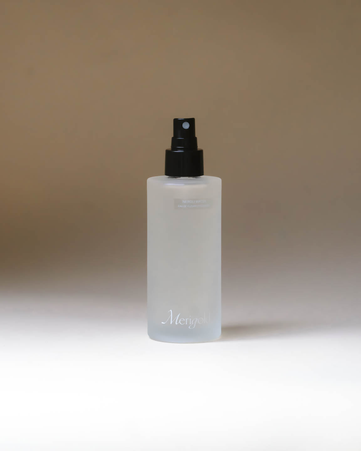 Clear spray bottle with Merigold logo in white on bottom of the bottle, black mister nozzle on a beige background