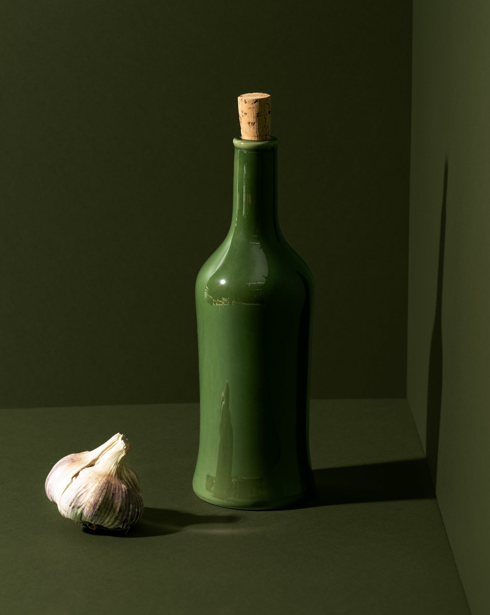 A white ceramic olive oil dispenser with a cork stopper, displayed next to a garlic bulb, on a neutral background.