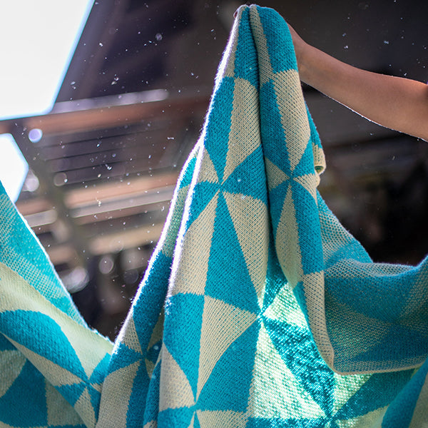 Teal and aqua blue patterned cotton throw blanket held up by a person against the sun.