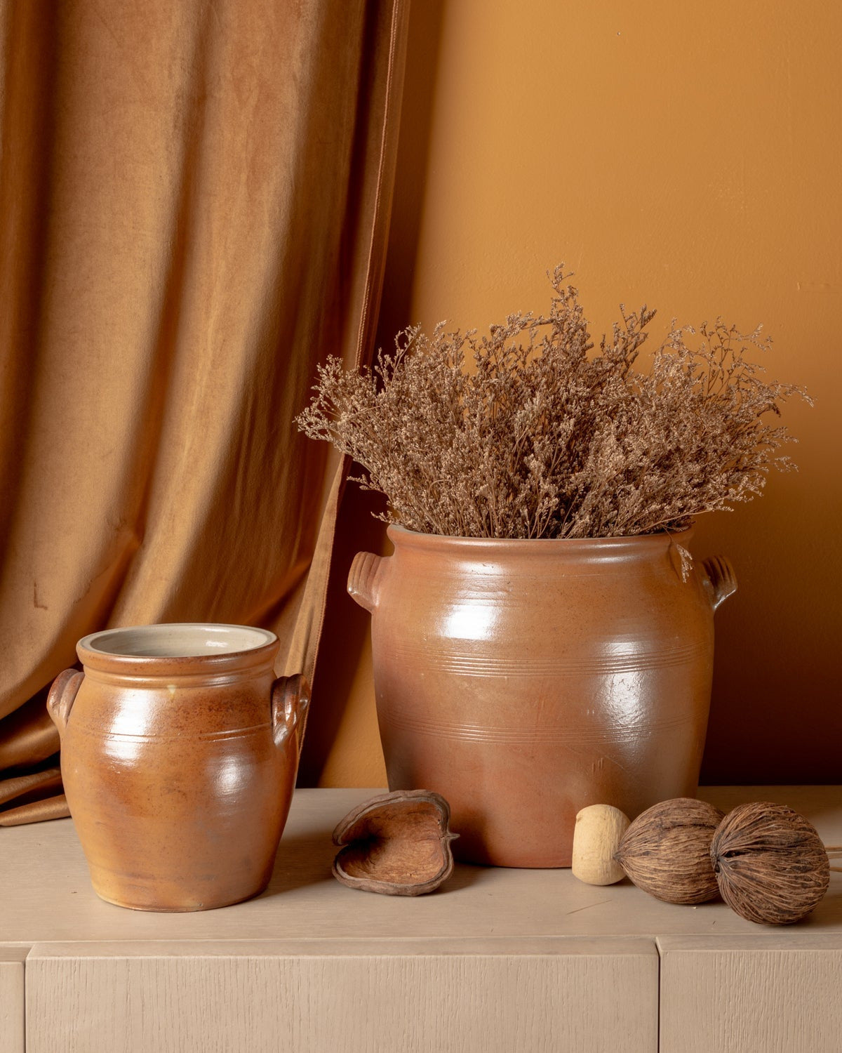 Two terracotta pots with dried plants and wooden elements on a beige surface against a brown curtain.