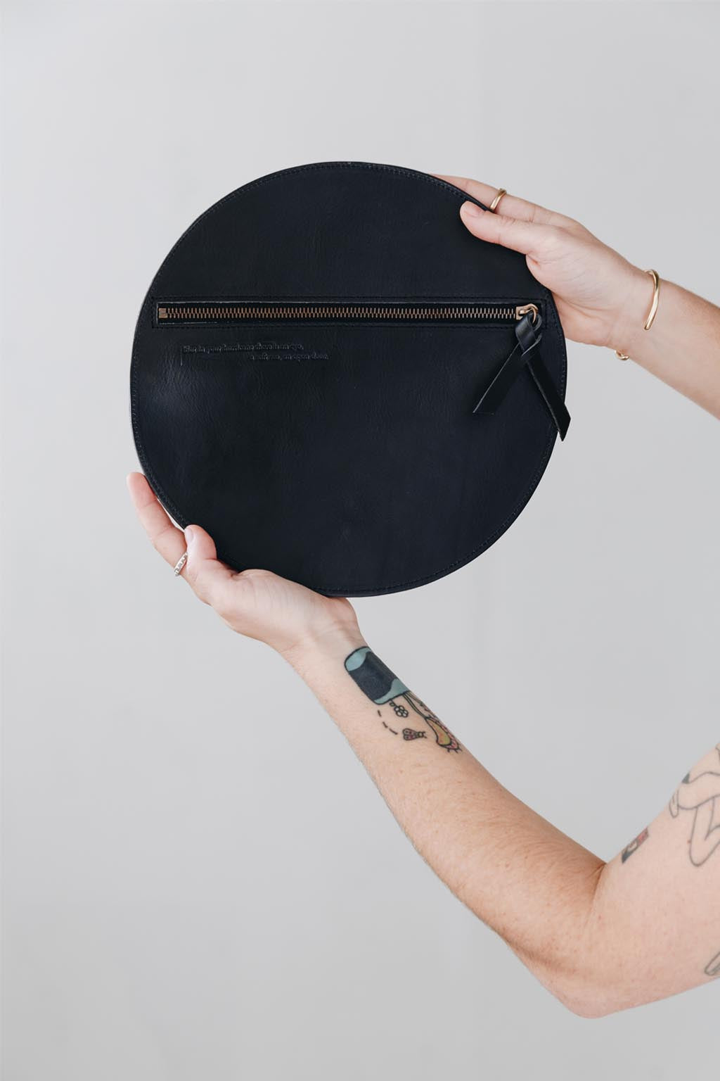 A woman's arms are visible, holding a circular black leather clutch with the reverse side and zipper visible. The zipper includes a matching zipper pull.