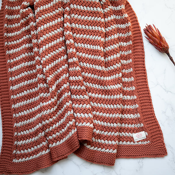 Rest rust coloured knitted throw blanket with a zigzag pattern on a white surface with an orange plant.
