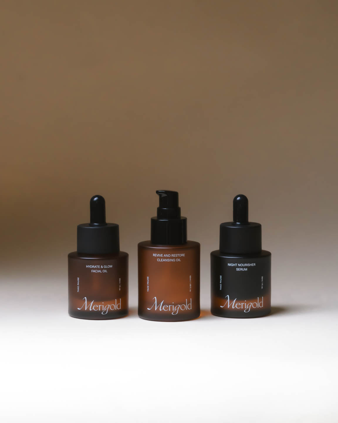 Merigold Oil Trio with Revive and Restore Cleansing Oil, Hydrate and Glow Facial Oil, and Night Nourisher Serum in amber glass bottles on minimal backdrop.