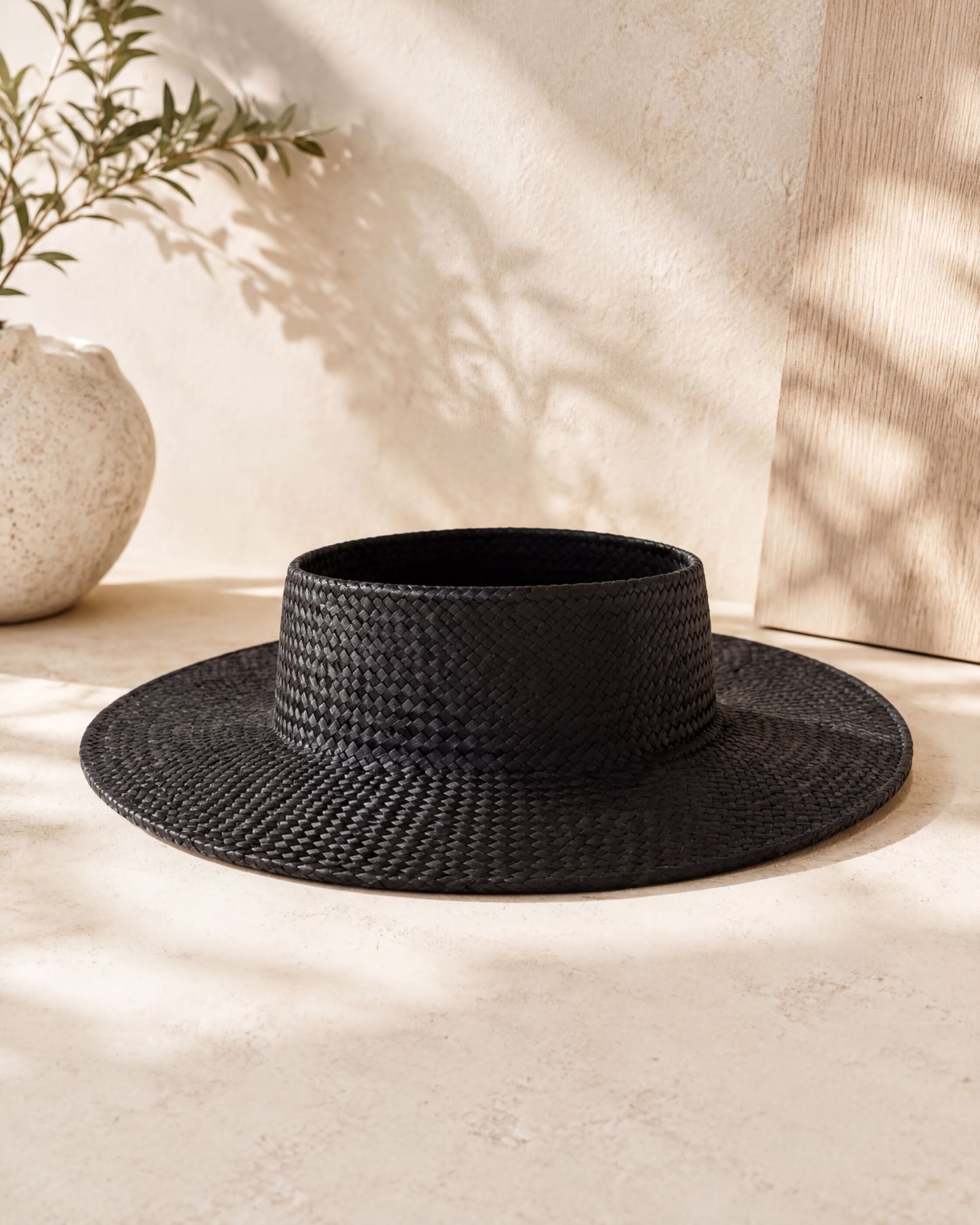 A black woven hat sits on a light-colored surface, with a white vase and a plant in the background.