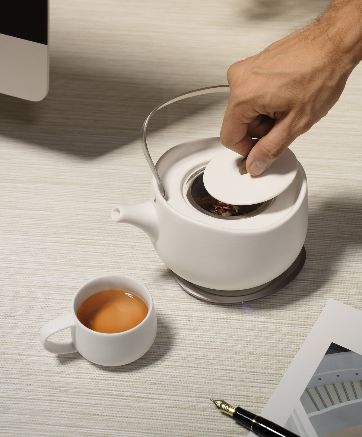 Person placing lid on Leiph self-heating teapot heating on OHOM Pad, with brewed teapot beside it
