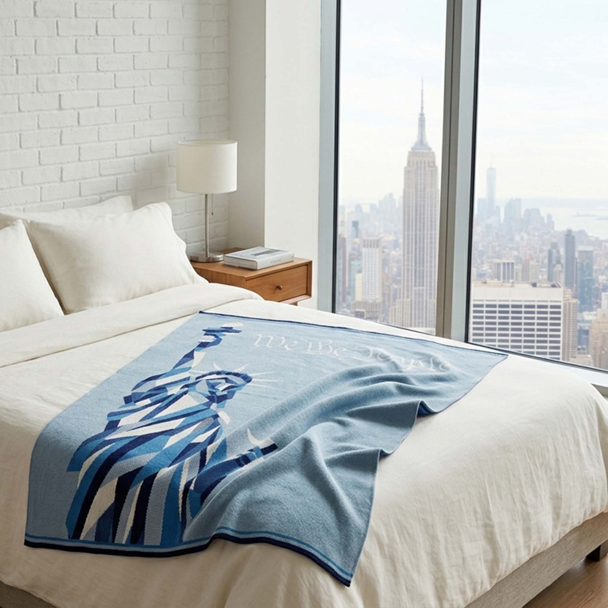 A neatly made bed is adorned with the in2green Statue of Liberty | We the People - Cotton Sustainable Knit Modern Throw Blanket, facing large windows overlooking the Empire State Building and New York City skyline.
